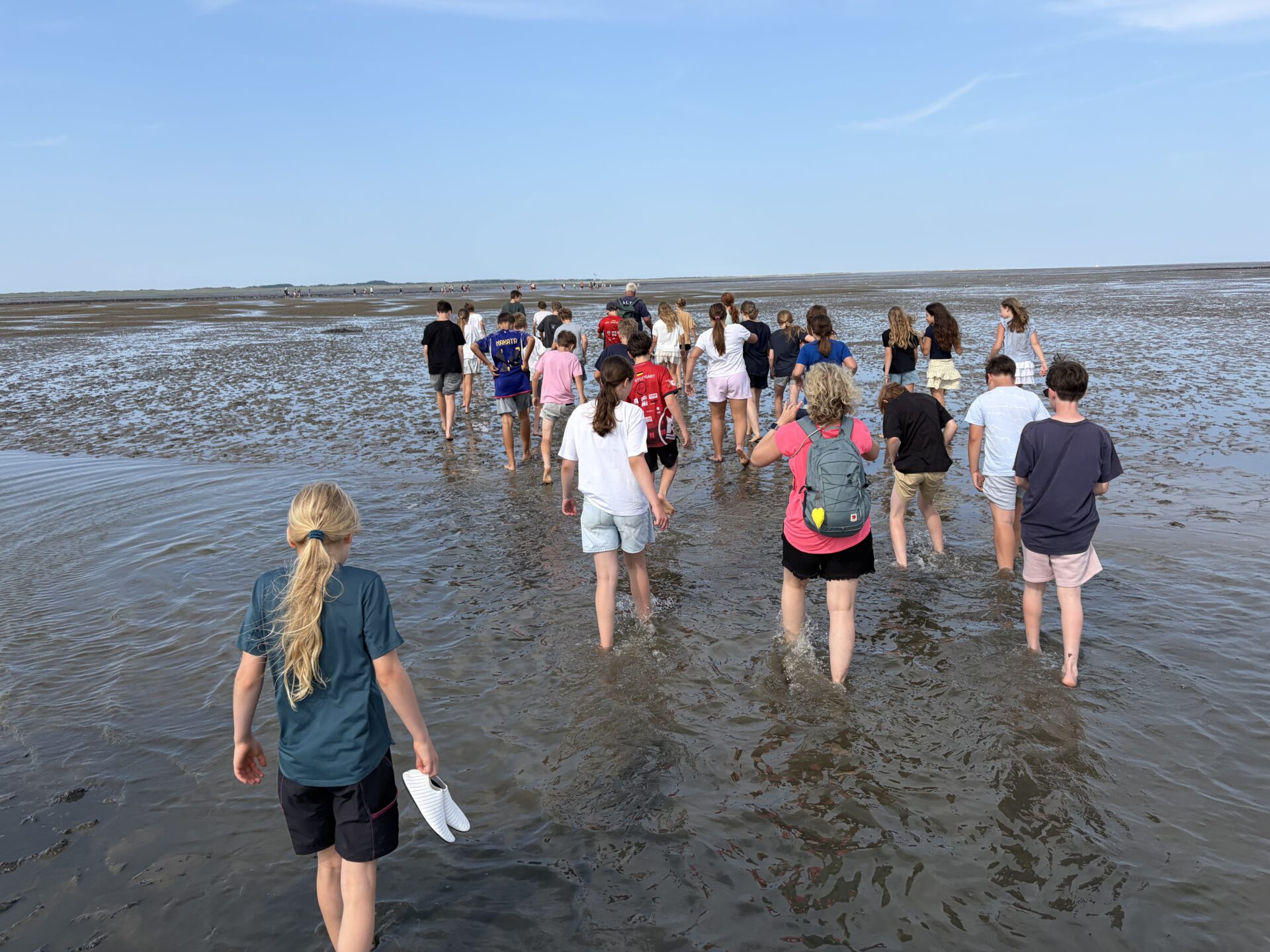 Eine Gruppe von Menschen, hauptsächlich Kinder, läuft barfuß durch seichtes Wasser an einem Sandstrand unter blauem Himmel. Einige halten ihre Schuhe in der Hand, und eine Person trägt einen roten Rucksack.