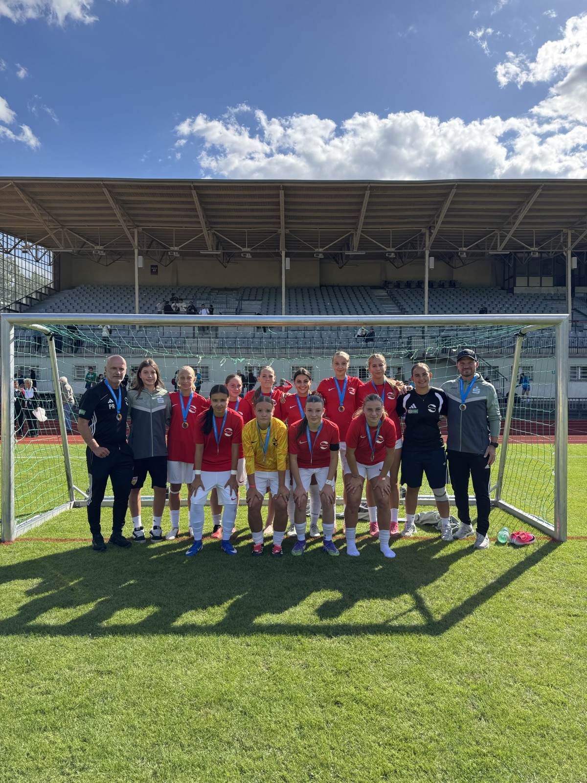 Eine Fußballmannschaft, die Medaillen trägt, posiert an einem sonnigen Tag vor einem Tor, während an beiden Enden Trainer stehen. Im Hintergrund ist unter einem blauen Himmel mit Wolken ein leeres Stadion mit Sitzplätzen zu sehen.