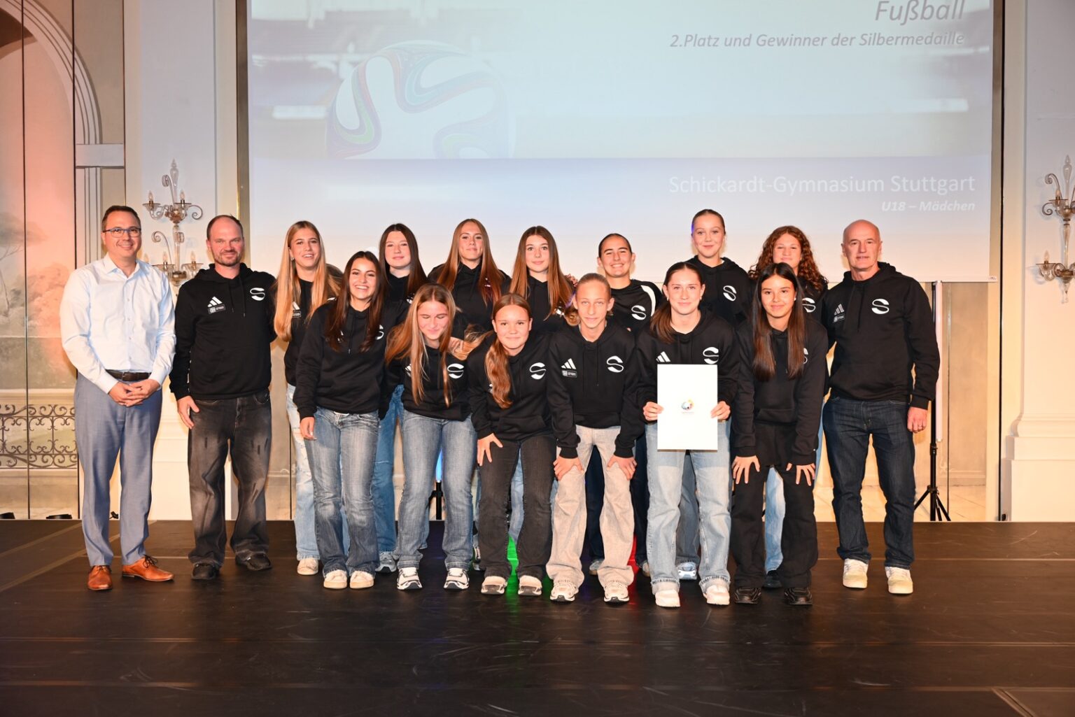 Eine Gruppe von Mädchen im Teenageralter und zwei Trainer stehen auf einer Bühne vor einer Leinwand, auf der sie als die Mädchenfußballmannschaft des Schickhardt-Gymnasiums Stuttgart ausgewiesen sind.