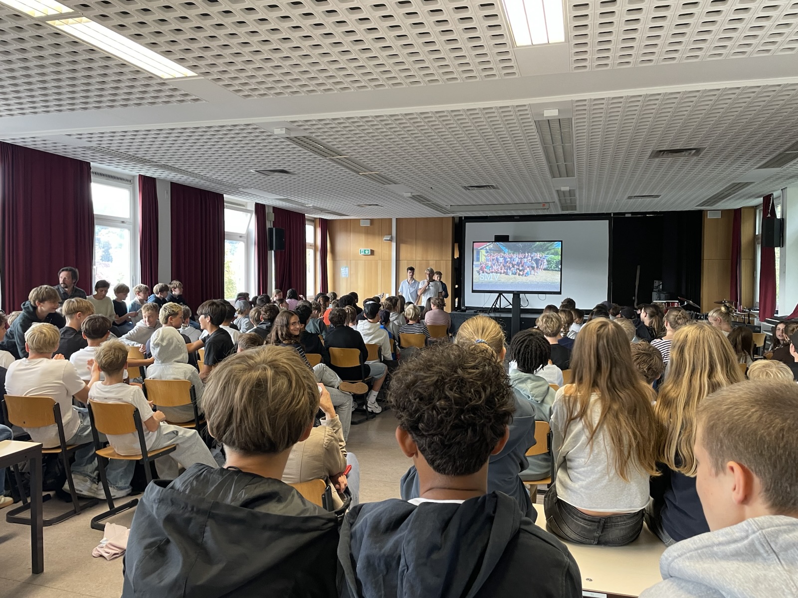 Eine Gruppe von Studierenden sitzt in einem Hörsaal und hört zwei Rednern zu, die neben einer großen Leinwand stehen, auf der ein Gruppenfoto zu sehen ist.